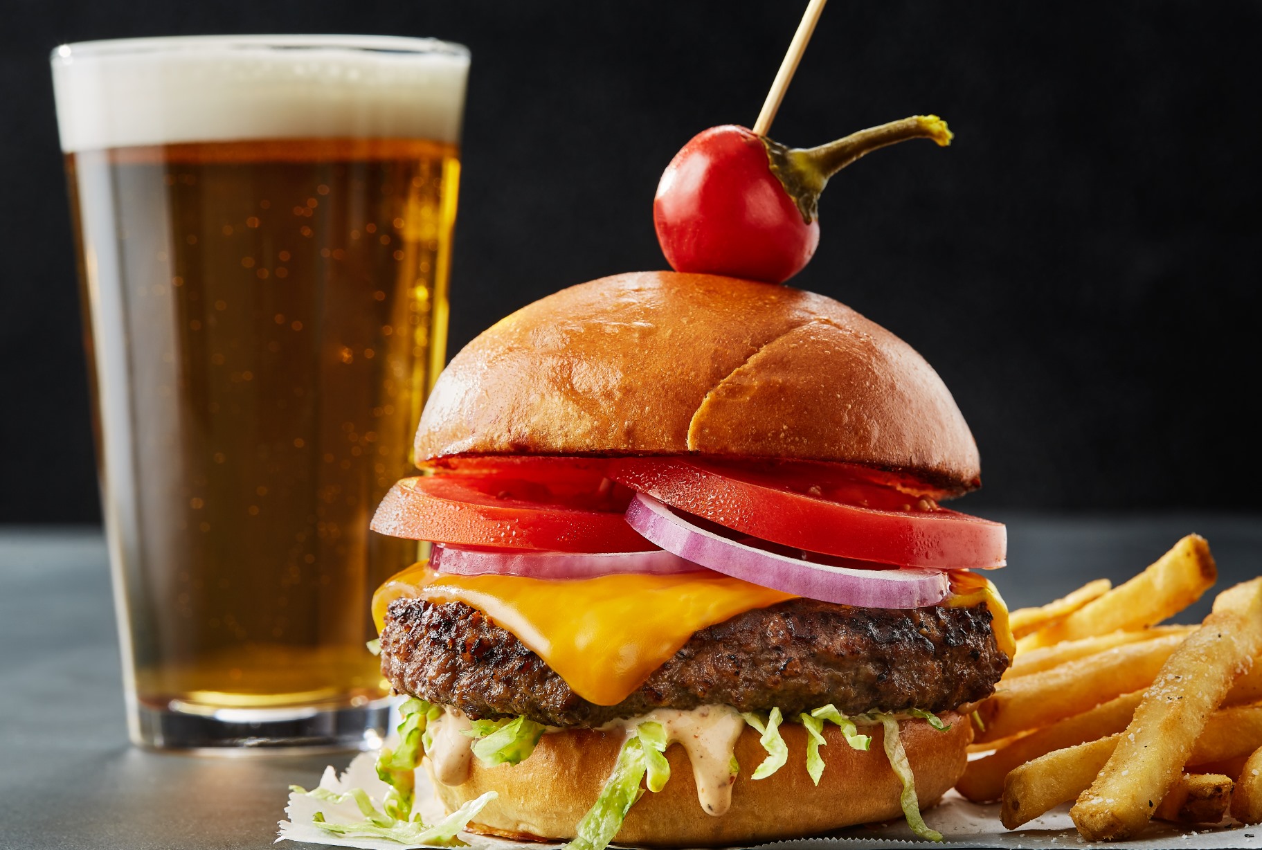 A cheeseburger with onions and tomatoes and a cherry tomato on top, along with French fries and a pint of beer from CRAVE