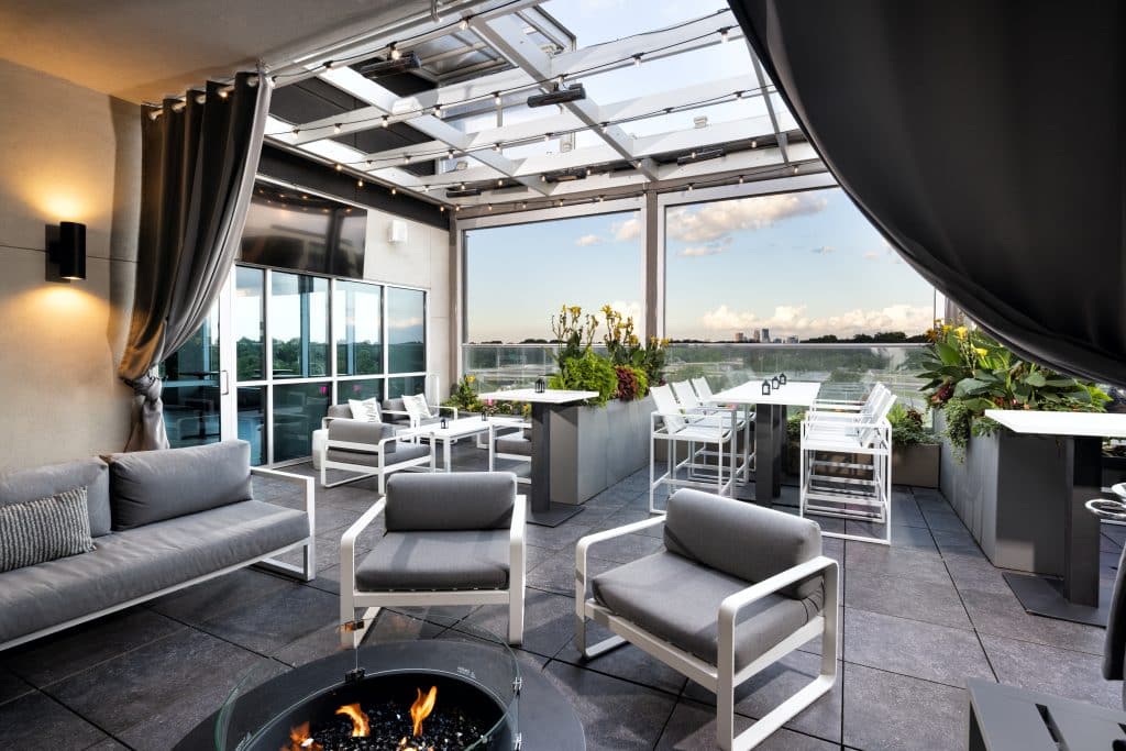 A photo taken from a cabana at REVEAL Rooftop Bar in St. Louis Park showing a fire pit, ample seating and a gorgeous skyline view