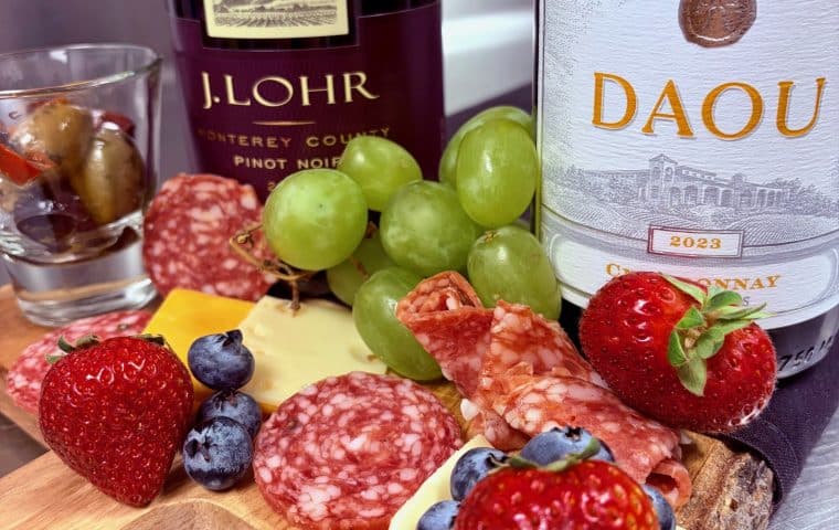 A photo of two bottles of wine and a charcuterie board with strawberries, cheese, grapes, salami and some olives to promote the tasting event at Three One Six