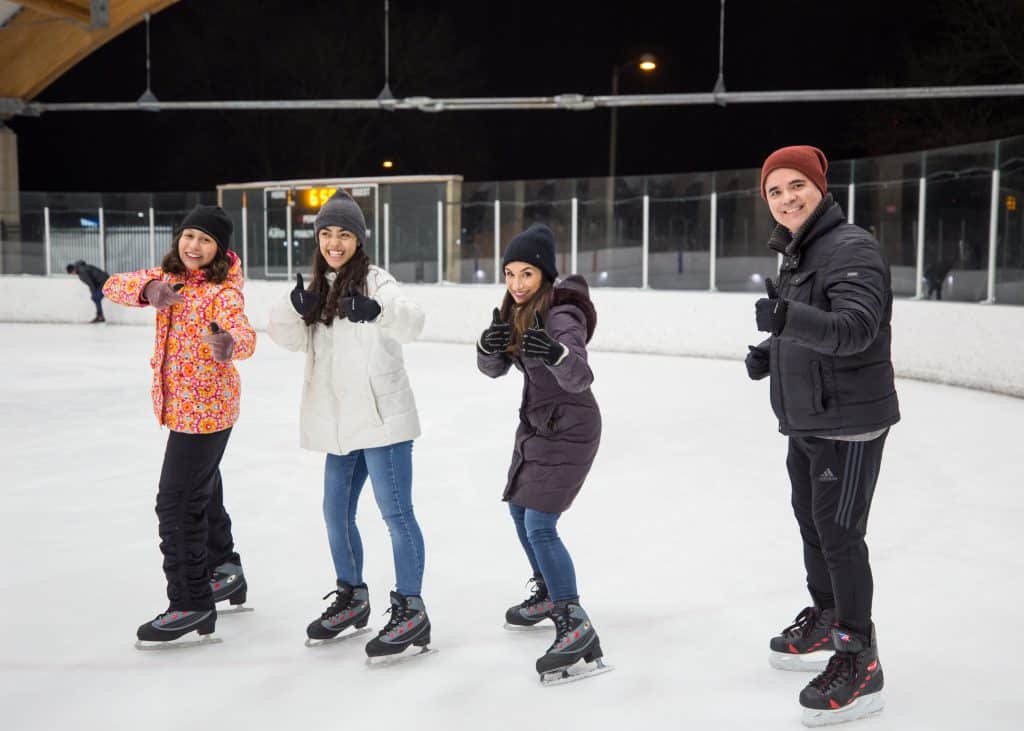 A mom, dad and two teenage daugthers ice skating at The ROC in St. Louis Park, smiling and pointing at the camera