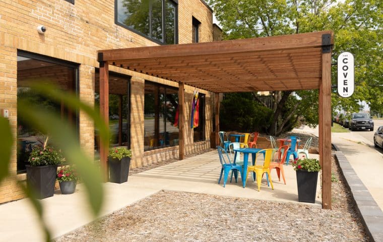 Exterior view of the building The Coven is in, showing a pergola covered patio with colorful tables and chairs
