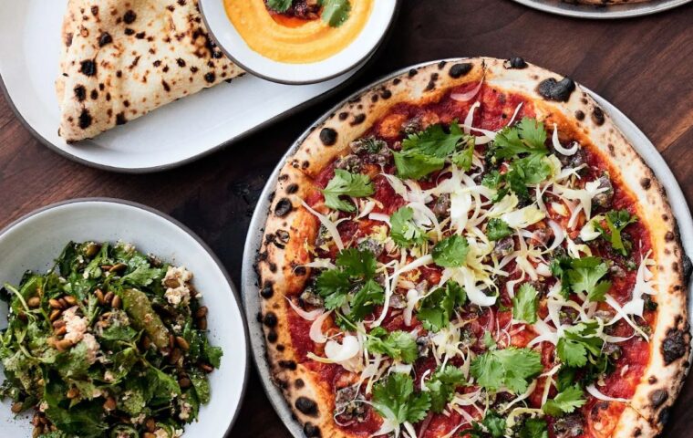 Photo of a wood-fired pizza next to a salad and a dish with pita bread and a dip