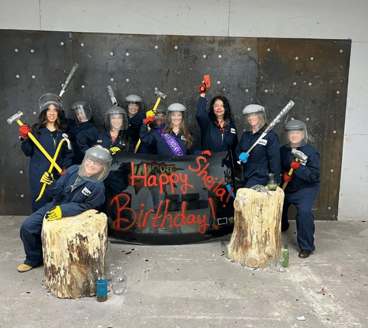 A group of women wearing coveralls and face shields holding sledgehammers next to a car windshield at iSMASH in St. Louis Park
